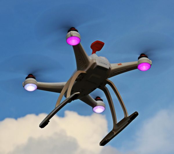 A quadcopter drone with propellers and lights flying high against a blue sky with clouds.